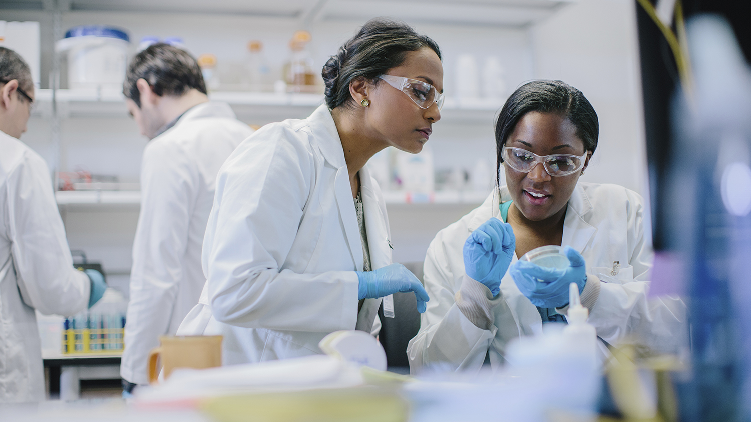 Two scientists in a lab are wearing white coats, gloves, and safety goggles. They are discussing data. Another scientist works in the background. The lab is filled with equipment and supplies.