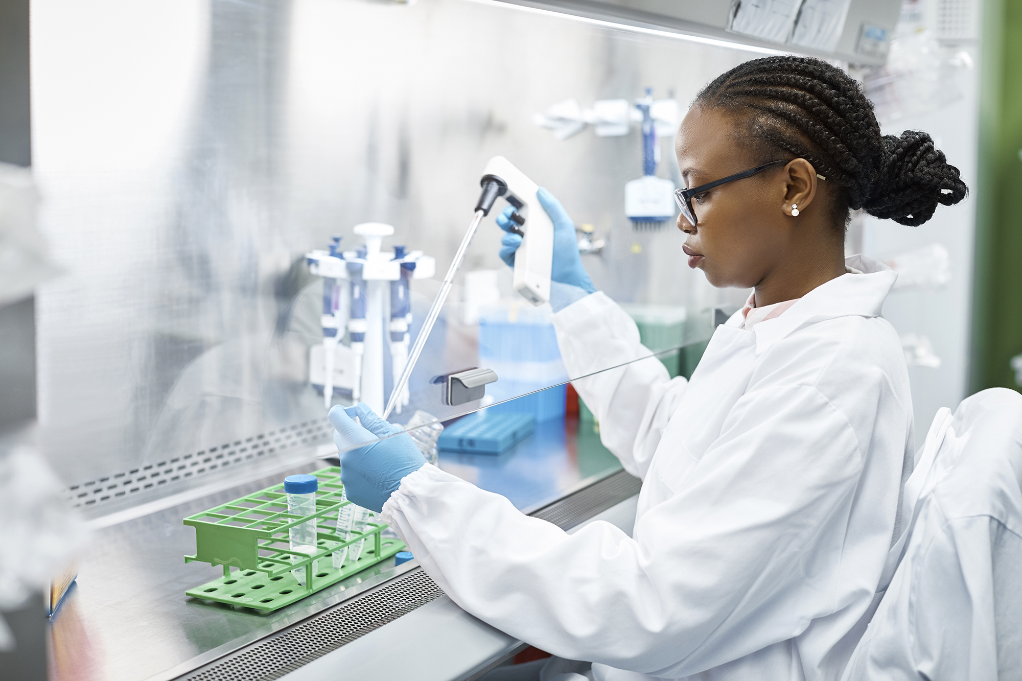 A female scientist in a white lab coat and blue gloves uses a pipette to transfer liquid into a test tube at a laboratory workstation with various equipment.
