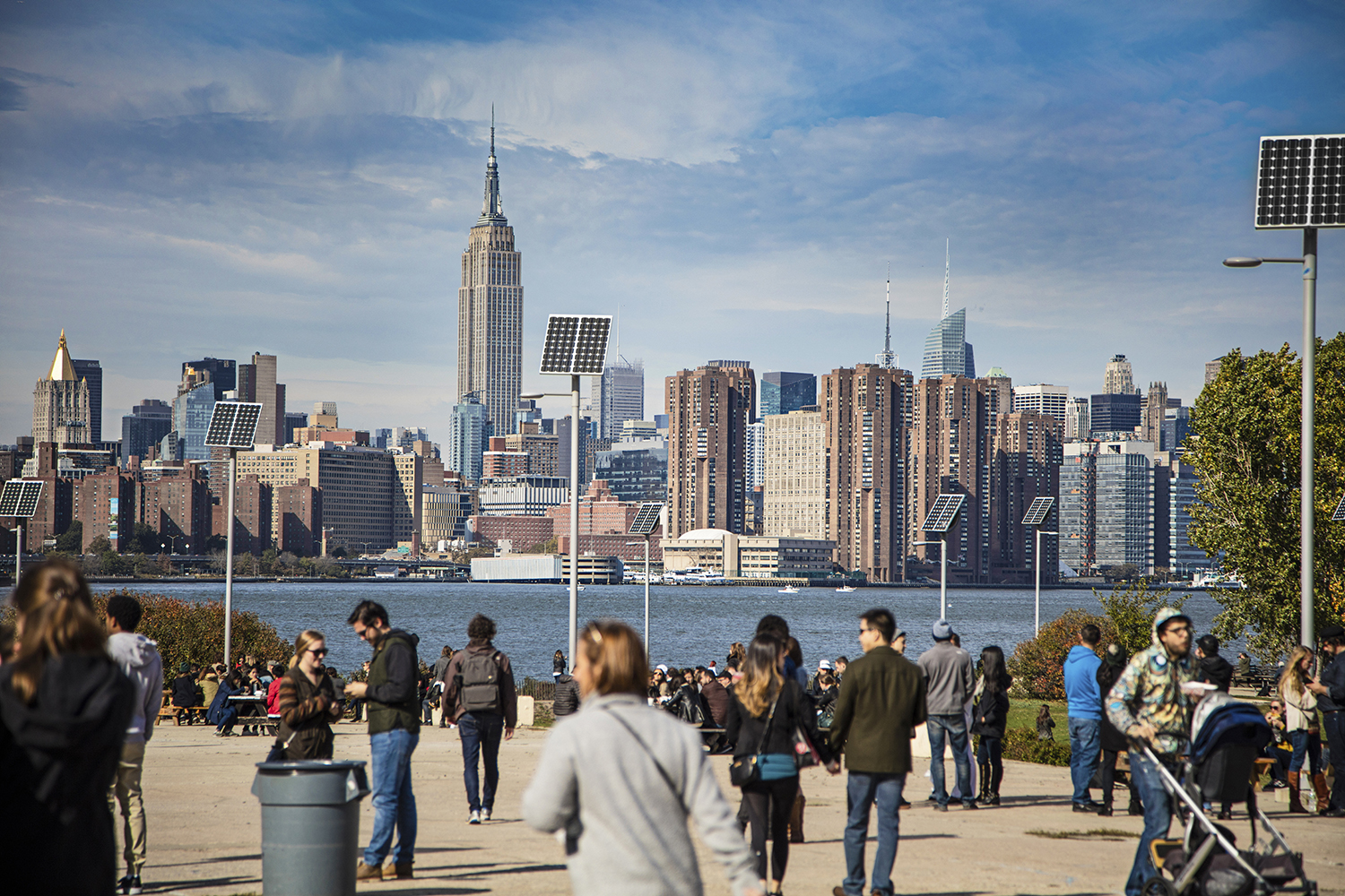 People on a promenade with solar panels and buildings in background. View of Manhattan is against sky.
