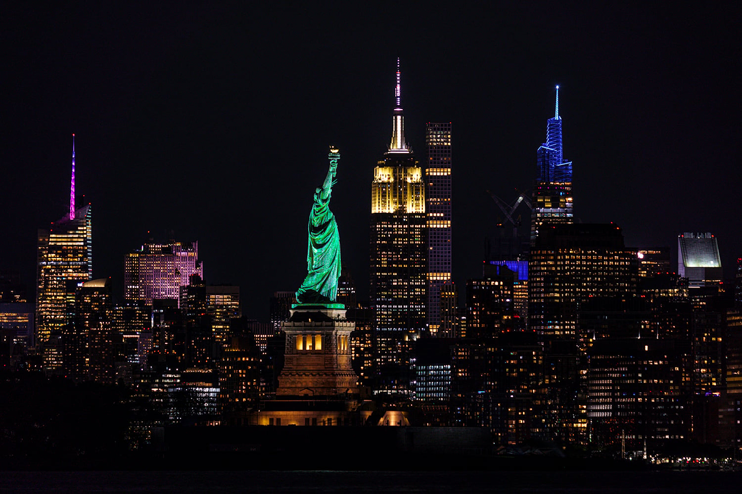 Night view of the New York City skyline featuring the illuminated Statue of Liberty in the foreground and skyscrapers in the background.