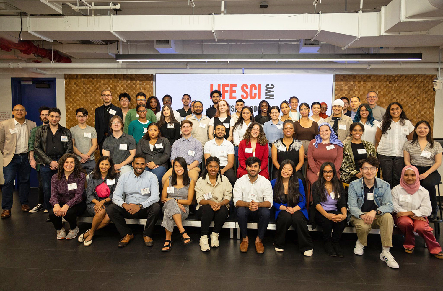 A diverse group of men and women smiling and posing for a group photo in a conference room. They are standing or sitting in front of a screen displaying Life Sci NYC. There are approximately 50 people in the image.