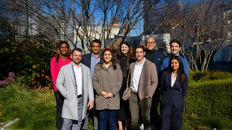 The LifeSci NYC team, a group of nine people, stands outdoors on a paved path with greenery and trees in the background, under clear skies.