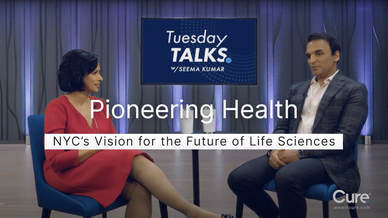 A woman in a red dress and a man in a suit sit on chairs facing each other in a studio, with a Tuesday Talks sign behind them. The text reads Pioneering Health: NYCs Vision for the Future of Life Sciences. The Cure logo is in the corner.