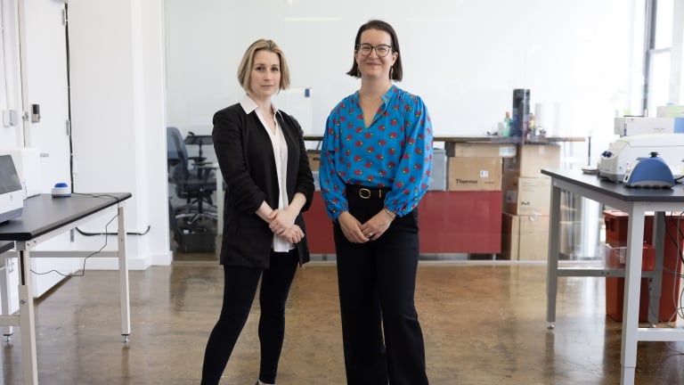 Two women, Cecilia Kushner and Beck Brachman, stand side by side in a modern, bright laboratory. One wears a black suit and heels, the other a blue blouse with a red pattern and black pants. Lab equipment and workstations are visible in the background. Two women, Cecilia Kushner and Beck Brachman, stand side by side in a modern, bright laboratory. One wears a black suit and heels, the other a blue blouse with a red pattern and black pants. Lab equipment and workstations are visible in the background.