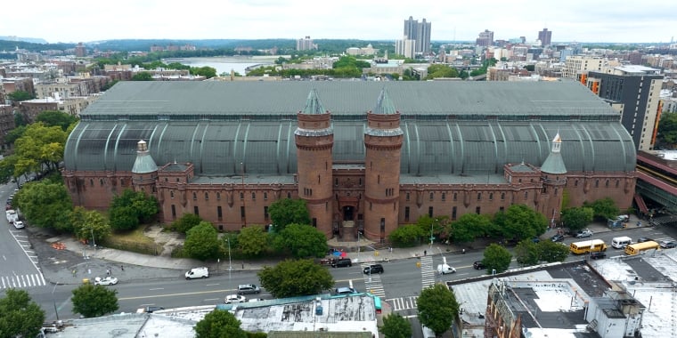 Aerial view of Kingsbridge Armory building with red brick walls and twin turrets, surrounded by NYC streets and greenery. The large, curved metal roof contrasts with the urban landscape, and several vehicles are visible on the roads nearby.