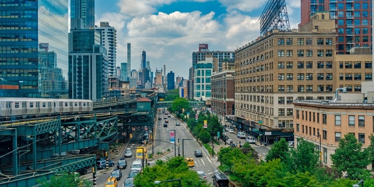 A busy urban street with cars and yellow cabs runs between tall buildings. A subway train travels on an elevated track to the left. The city skyline is visible in the background under a partly cloudy sky.