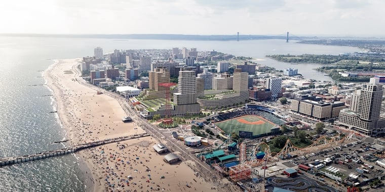 Aerial view of a Coney Island featuring a long sandy beach crowded with people, an amusement park with rides, a baseball stadium, and numerous buildings. The coastline curves with a distant bridge visible on the horizon.