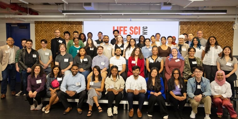 A diverse group of men and women smiling and posing for a group photo in a conference room. They are standing or sitting in front of a screen displaying Life Sci NYC. There are approximately 50 people in the image.