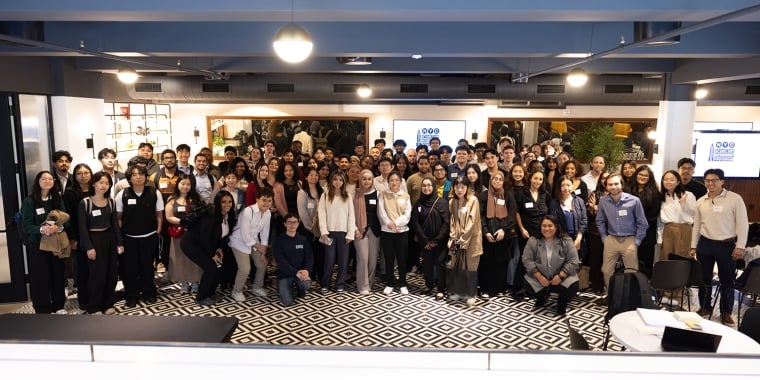 NYC Startup Internship program participants pose for a group photo indoors, many wearing name tags and business attire, standing on a black-and-white patterned floor in a spacious, modern room.