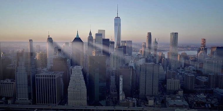 Aerial view of Lower Manhattan, New York City, at sunrise with sunlight streaming between tall skyscrapers including One World Trade Center, and the Hudson River in the background.