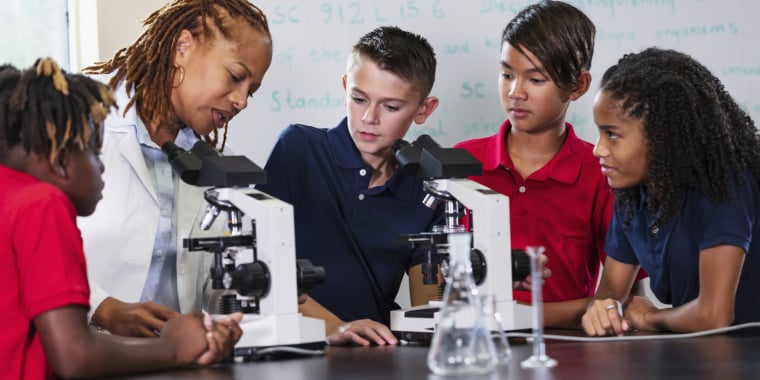 A teacher and four students gather around microscopes in a science classroom, examining slides and discussing their observations. The students look engaged and curious. Laboratory glassware is on the table.